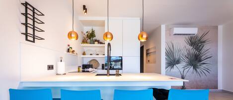 A modern kitchen with a central island, blue chairs, pendant lighting, and decorative plants.