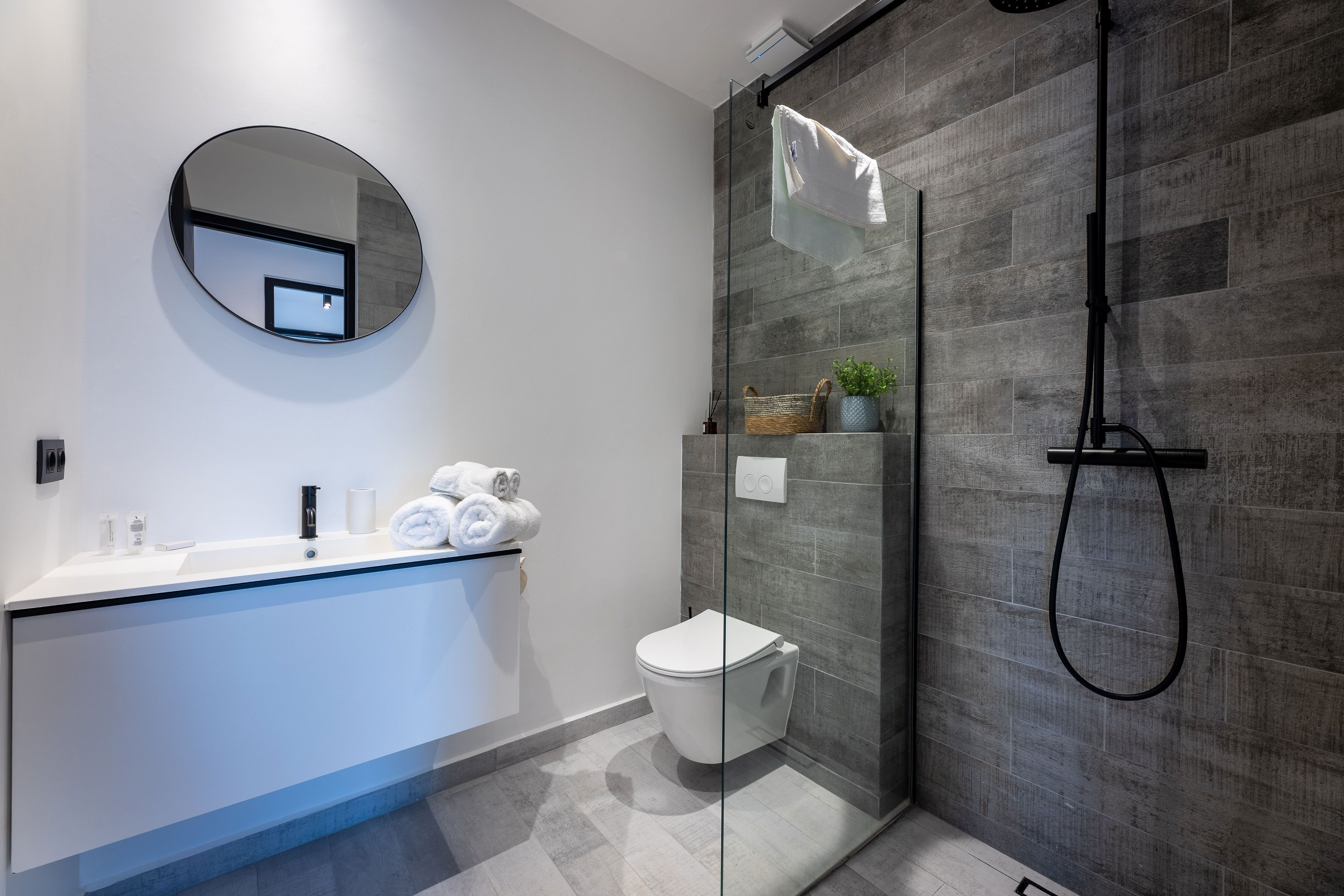 Un baño moderno con cabina de ducha de vidrio, un espejo redondo, un lavabo blanco y un inodoro.