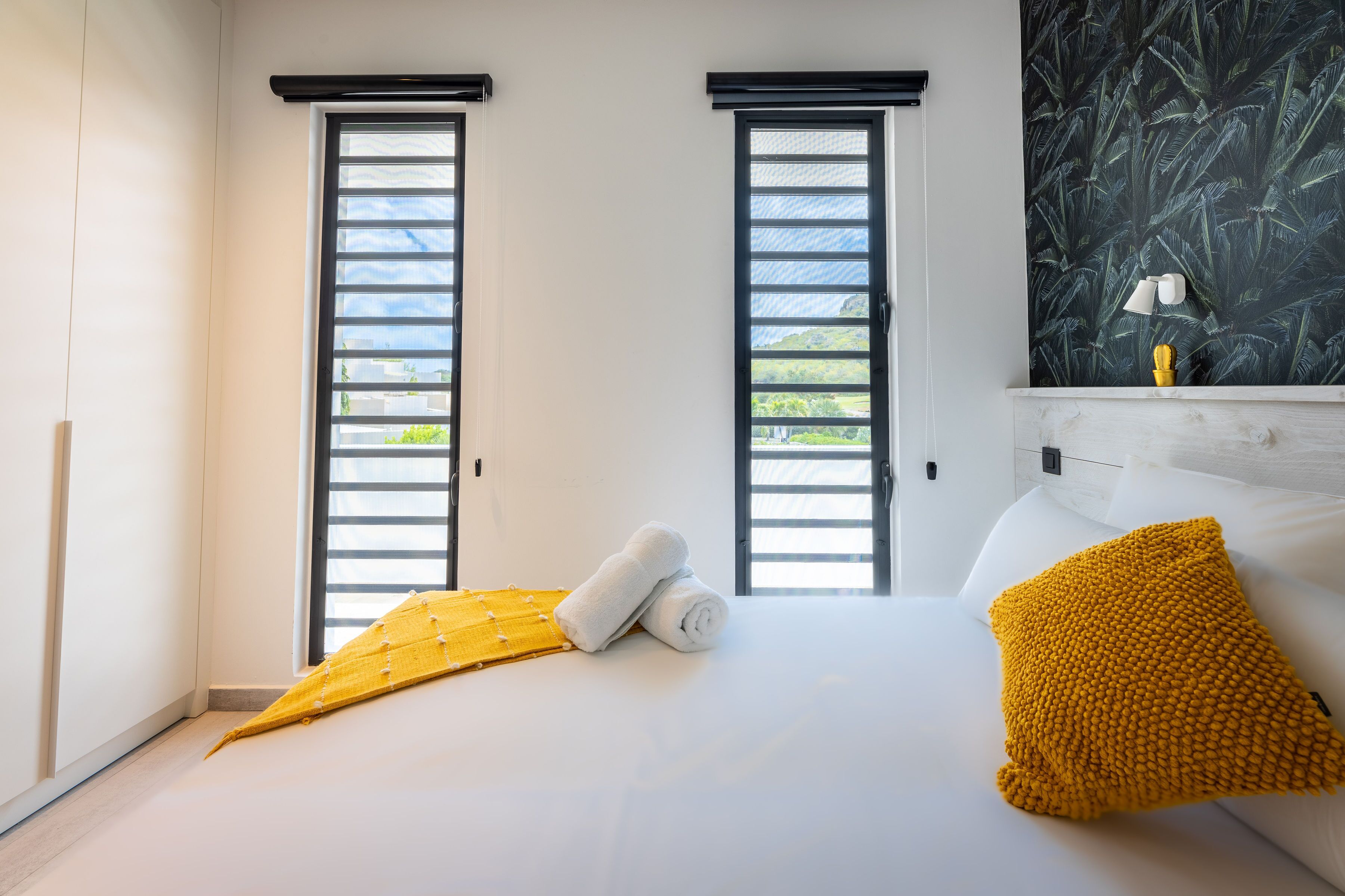Une chambre à coucher avec un lit, deux fenêtres équipées de stores, un plaid jaune, un coussin jaune et un mur orné d’un motif de feuilles.