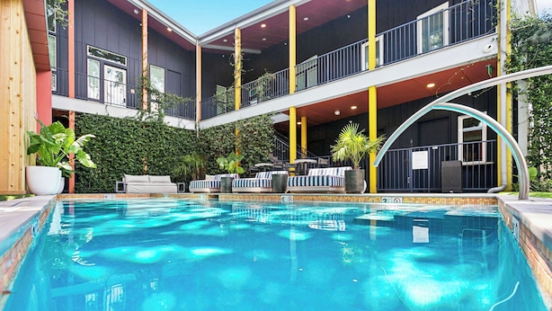 Une piscine dotée de chaises longues et d’un espace salon couvert, le tout entouré d’un bâtiment agrémenté de balcons et d’un mur végétal.
