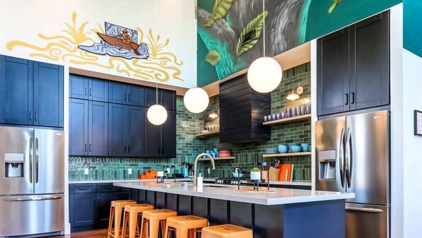 Una cocina moderna con armarios oscuros, una isla central con taburetes de bar, electrodomésticos de acero inoxidable y un mural en la pared con motivos de hojas verdes.