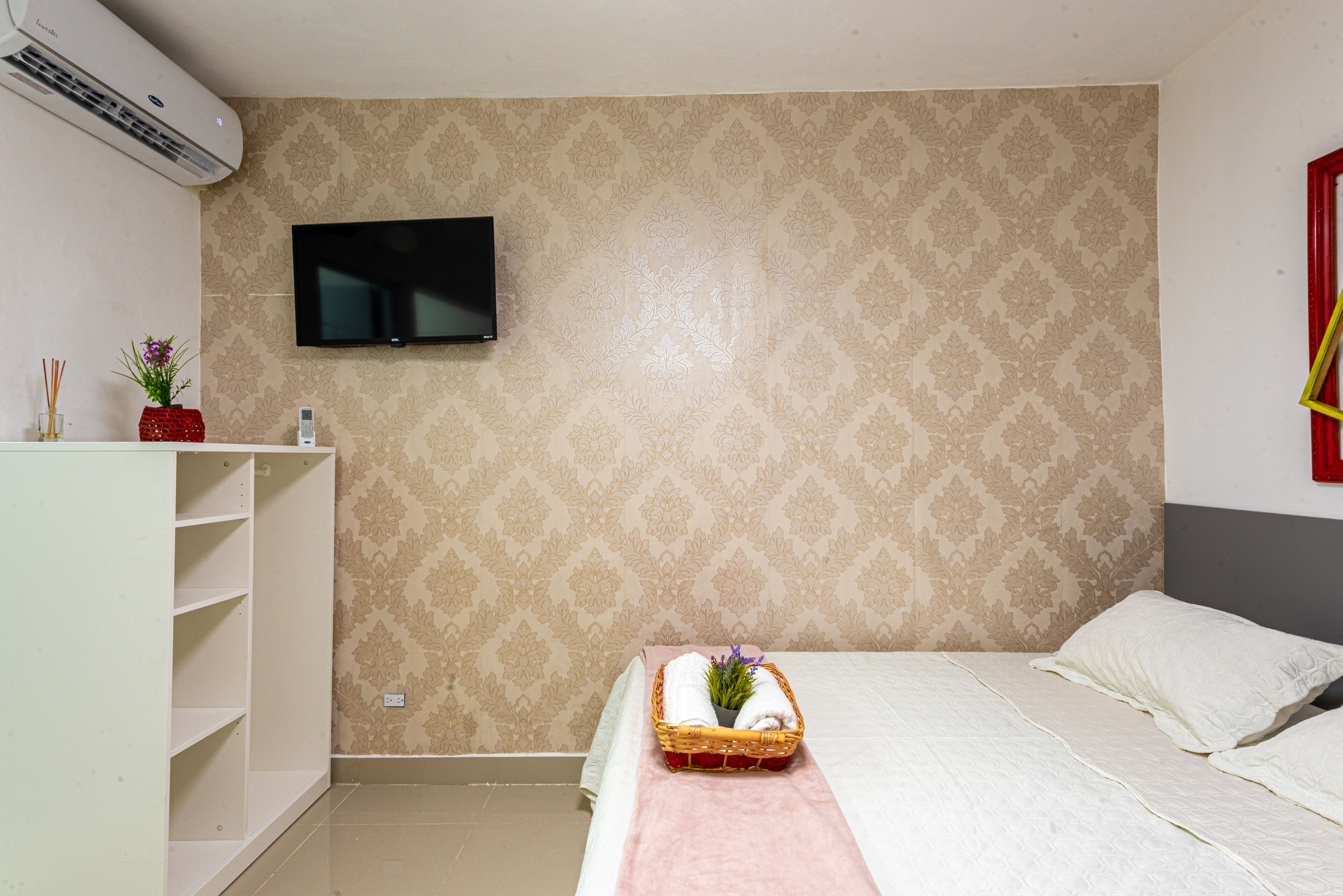 Een netjes ingerichte hotelkamer met een bed, een aan de muur bevestigde televisie, een plankenunit en een rode vaas met bloemen.