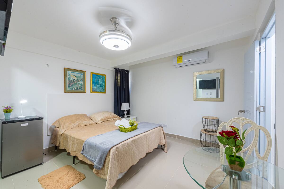 A hotel room with a bed, a desk, a refrigerator, a mirror, and a glass table with a vase of flowers.