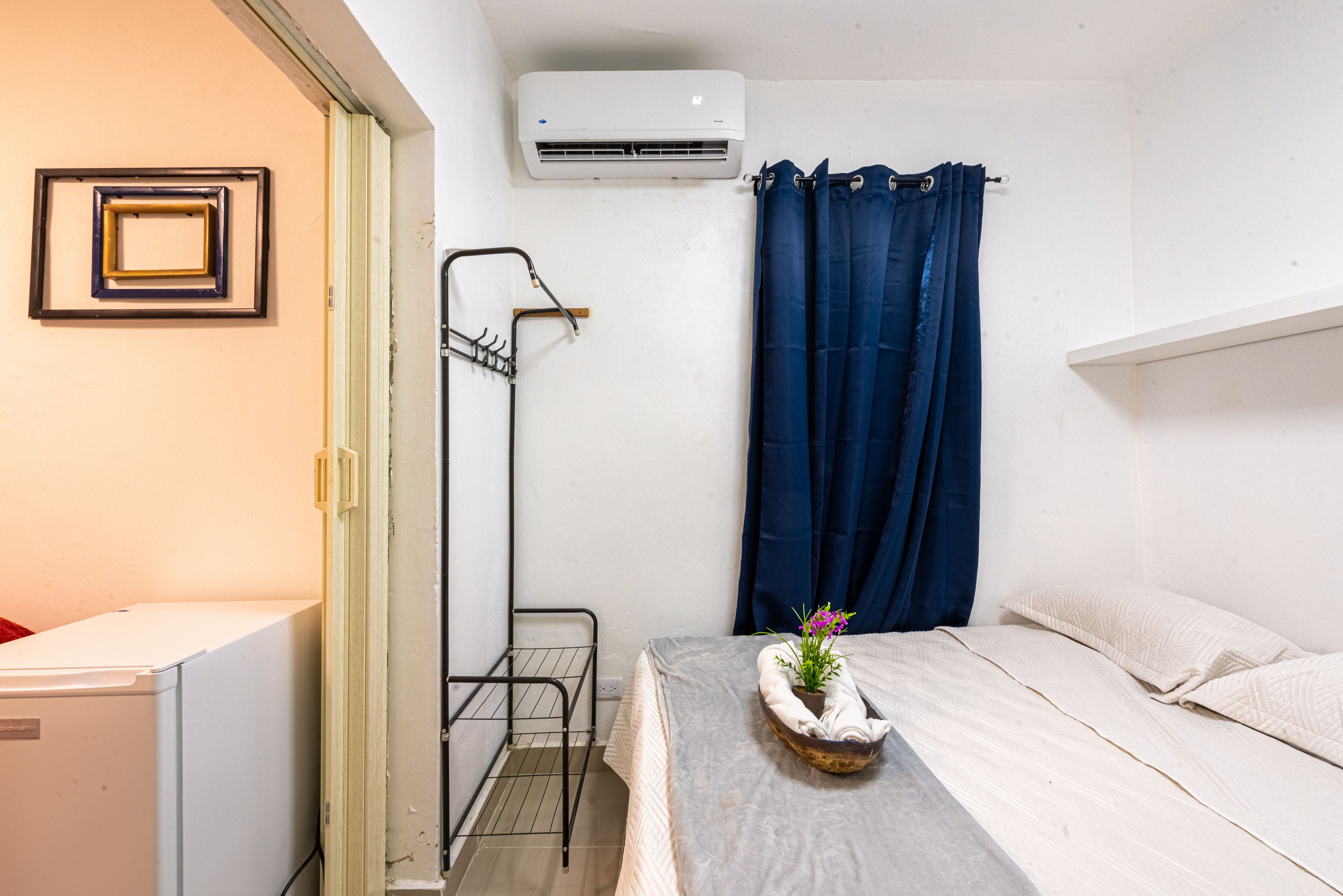 Een kleine kamer met een bed, een klein tafeltje met een plant, een witte airconditioner, een metalen rek en een blauw gordijn.