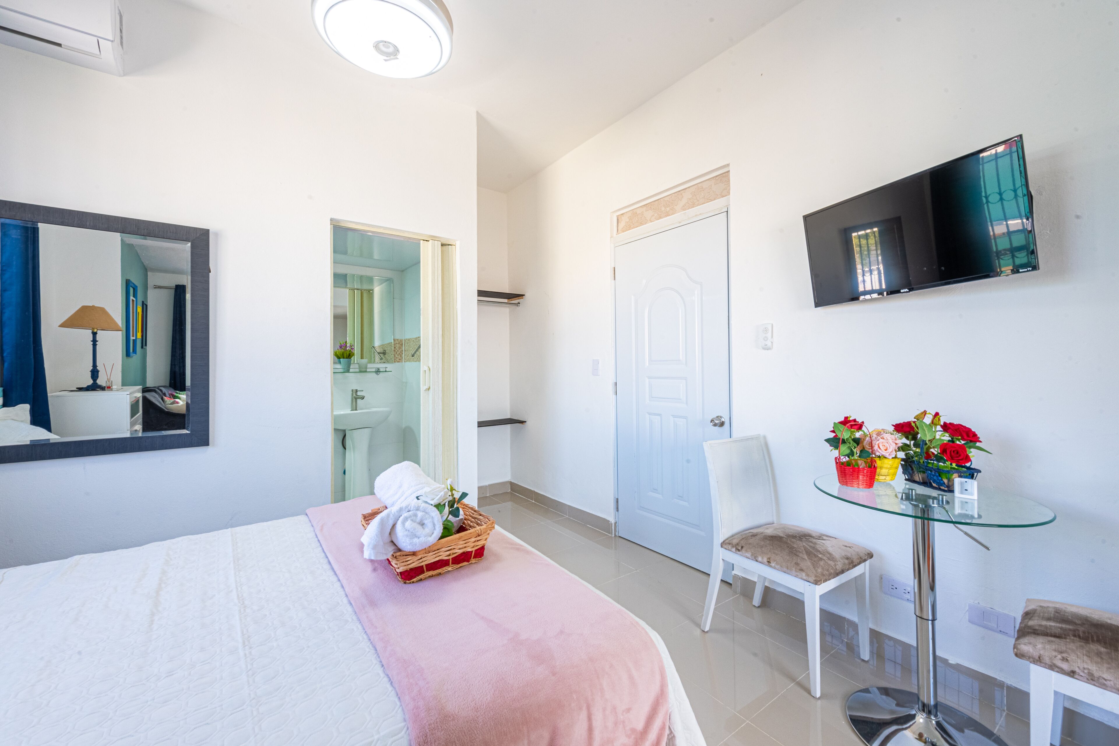 A hotel room with a bed, a glass table with flowers, a chair, a TV mounted on the wall, and a door.