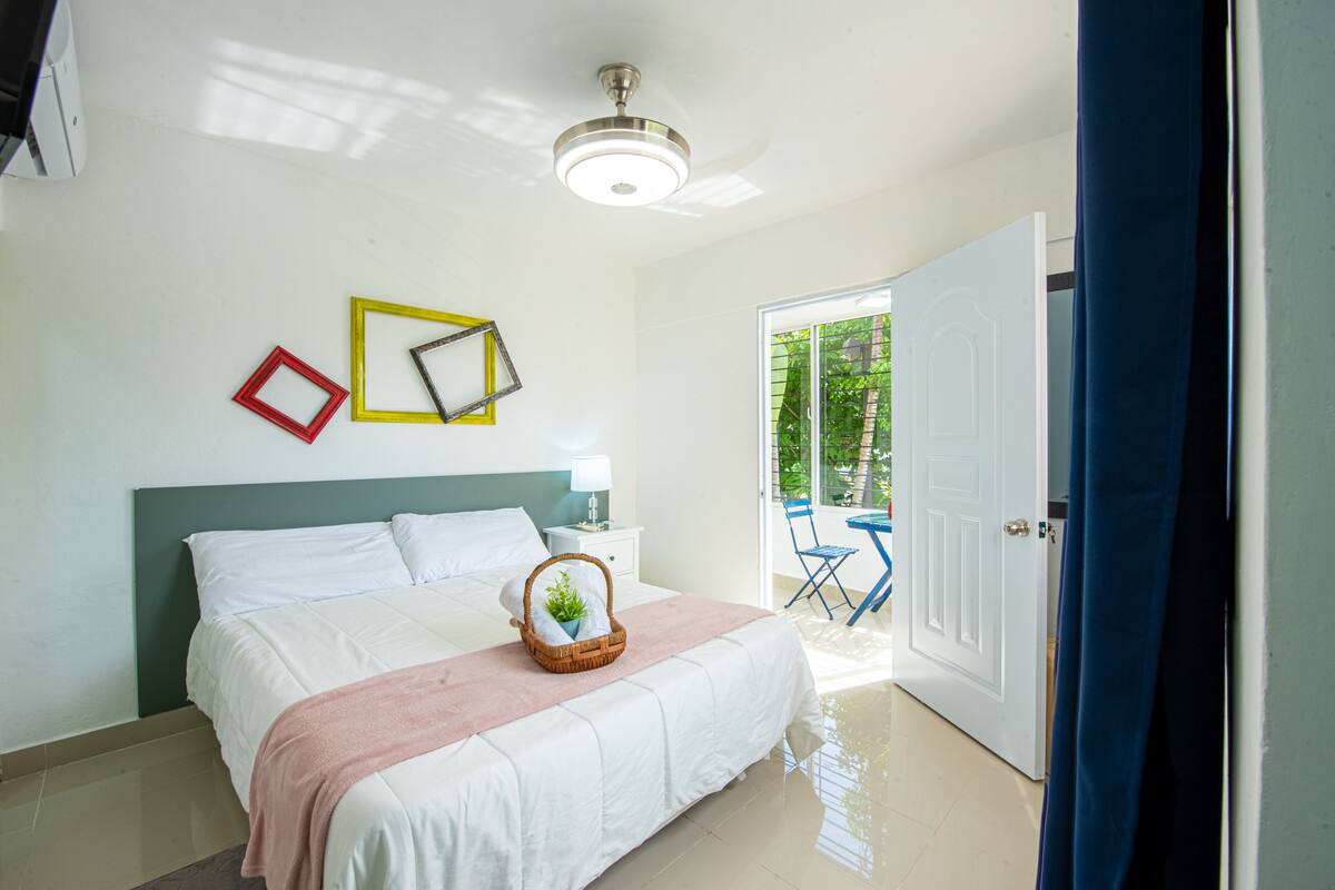 A bedroom with a bed, bedside table, lamp, and a view of the outdoors.