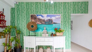 A room with a white desk, a large framed picture of a coastal landscape, bamboo wallpaper, potted plants, and a red decorative shelf. - Bamboo House (Santo Domingo)