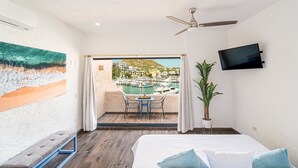 A hotel room with a bed, a ceiling fan, a TV, a balcony with a view of a marina and buildings, and a painting on the wall. - Newly Remodeled Condo In The Cabo Marina (Cabo San Lucas)