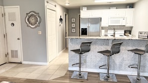 A modern kitchen with white cabinets, a stainless steel refrigerator, a microwave, and a coffee maker. There are three bar stools at the counter.