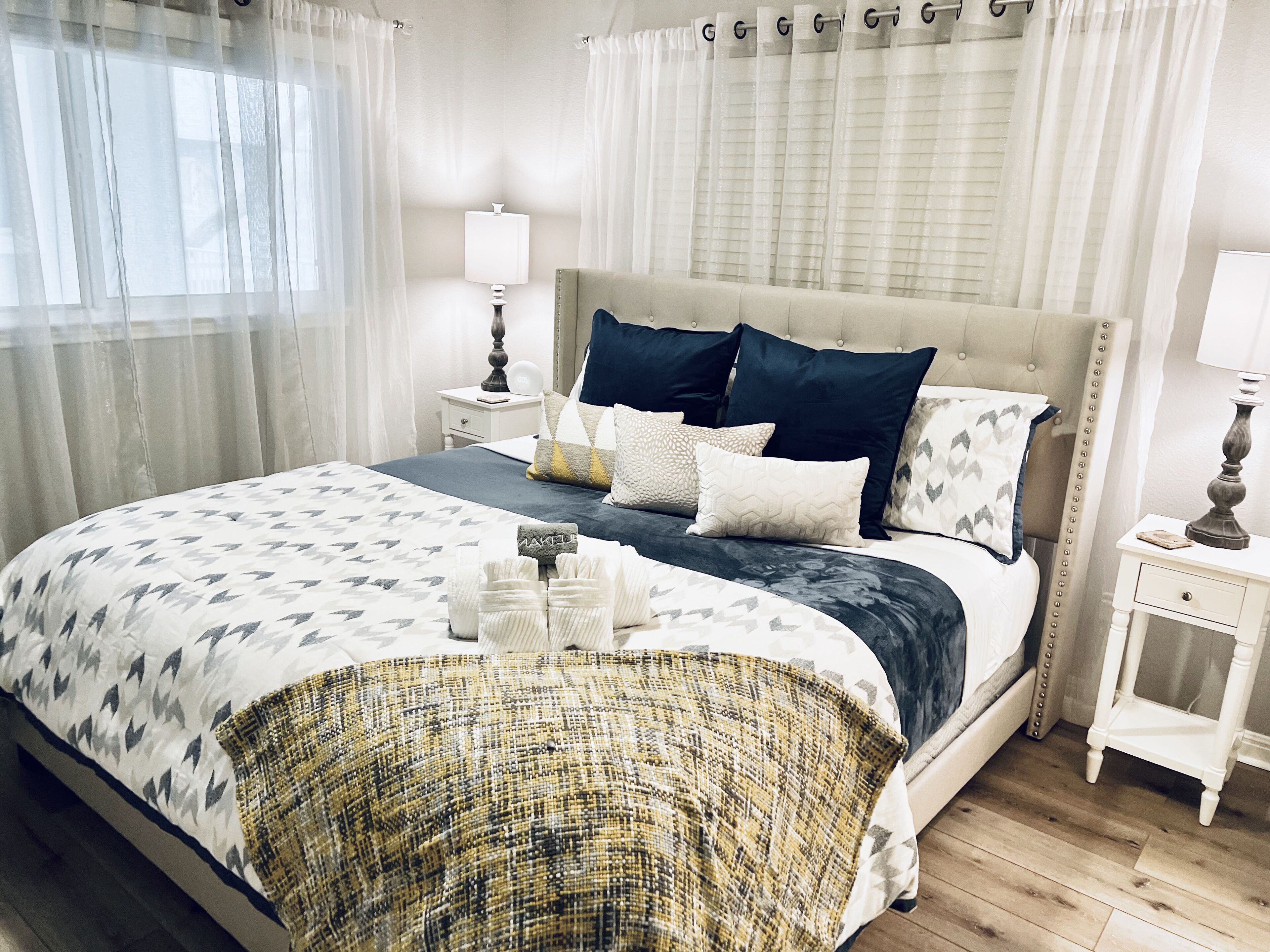 Quarto bem organizado com uma cama grande, mesa de cabeceira, luminária e janela com cortinas leves.