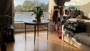 Een ruime woonkamer met een groot raam, een houten vloer, een ronde tafel met een plant, een bank met decoratieve kussens, en een kledingkast met daarin hangende kleding.