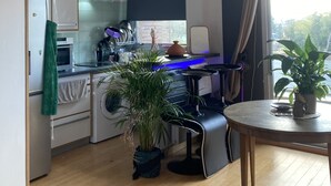 Een compacte keuken met een koelkast, oven en magnetron. Er is een eethoek met een tafel en stoelen, en een potplant.
