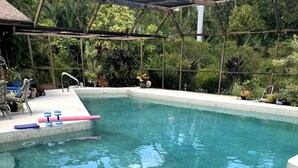 Una piscina cristalina con estructura de techo metálico, rodeada de abundante follaje.