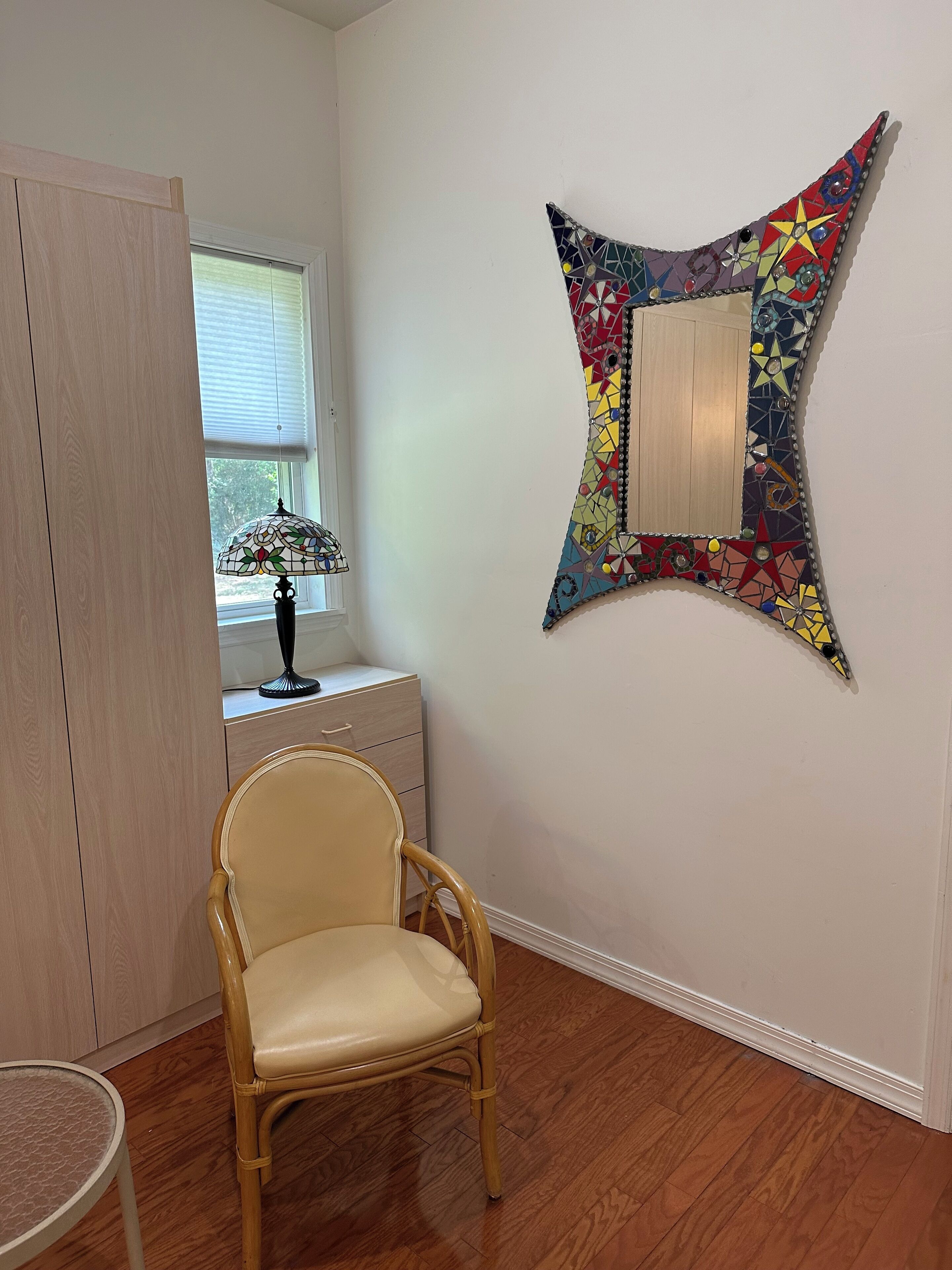 Ein Zimmer mit Holzboden, einem Sessel, einem Tisch, einem Fenster mit Jalousien, einer Lampe und einer farbenfrohen Wanddekoration.
