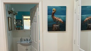 Un pasillo con una puerta blanca que da a un baño con lavamanos y armarios. Hay dos cuadros en la pared, uno de un pelícano y otro de una escena similar.