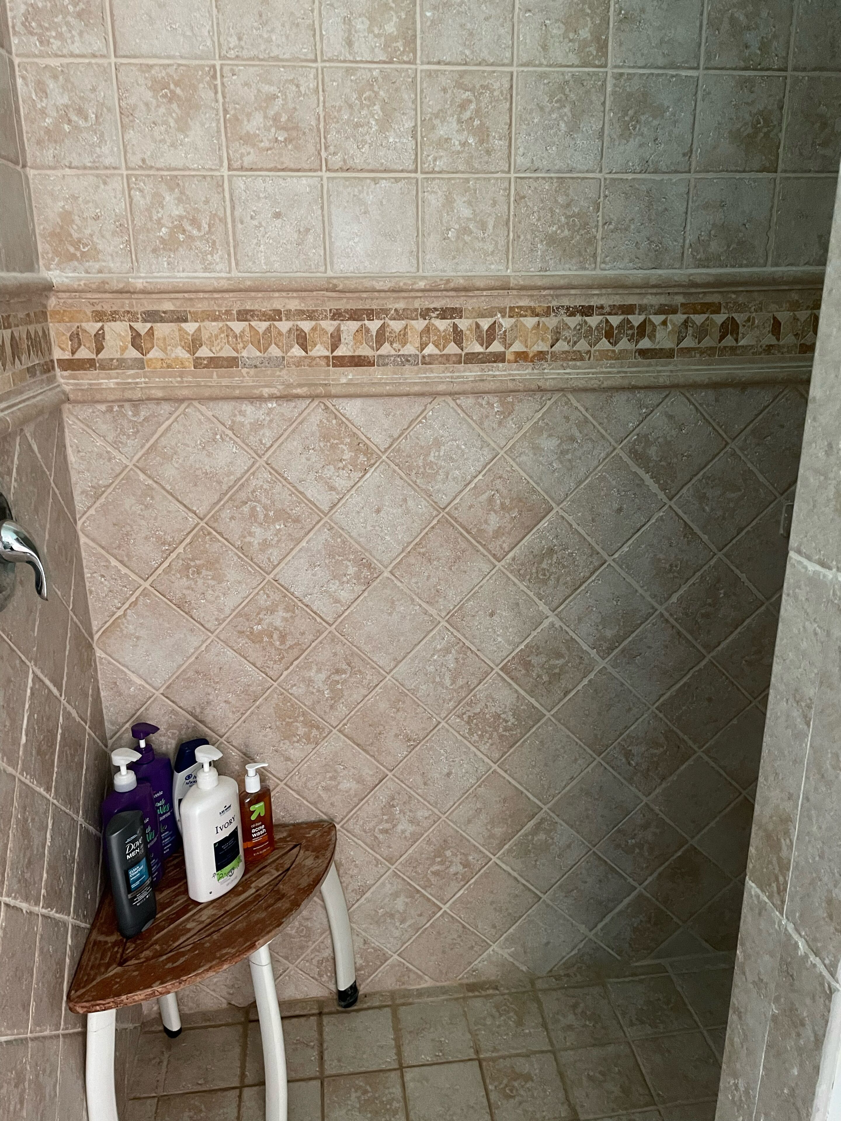 Une salle de bain avec des murs et un sol carrelés de couleur beige, une étagère en bois où sont rangés les articles de toilette, et un espace douche.