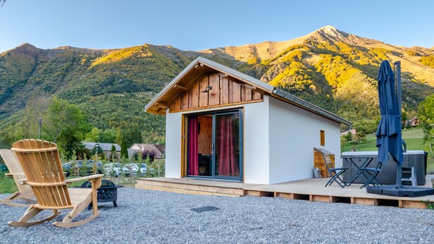 Eine kleine Holzhütte mit einer Schiebetür aus Glas, einer Terrasse mit Gartenmöbeln und Blick auf die Berge im Hintergrund.