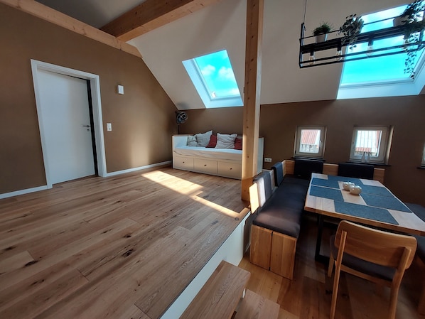 A spacious room with a loft bed, a dining area with a table and chairs, wooden flooring, and skylights allowing natural light.