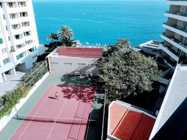 A tennis court with a red surface, surrounded by buildings and a view of the sea. - 2 Towers Sea View in Funchal Swimming Pool & Tennis (Funchal)
