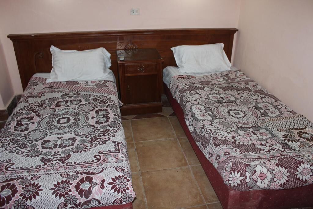 Una habitación con dos camas individuales, cada una con una colcha estampada y almohadas blancas. Entre las camas hay una mesita de noche de madera con un cajón pequeño y una lámpara. El piso es de baldosas y la pared es lisa, con un interruptor de luz sobre la mesita.