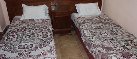 Una habitación con dos camas individuales, cada una con una colcha estampada y almohadas blancas. Entre las camas hay una mesita de noche de madera con un cajón pequeño y una lámpara. El piso es de baldosas y la pared es lisa, con un interruptor de luz sobre la mesita.