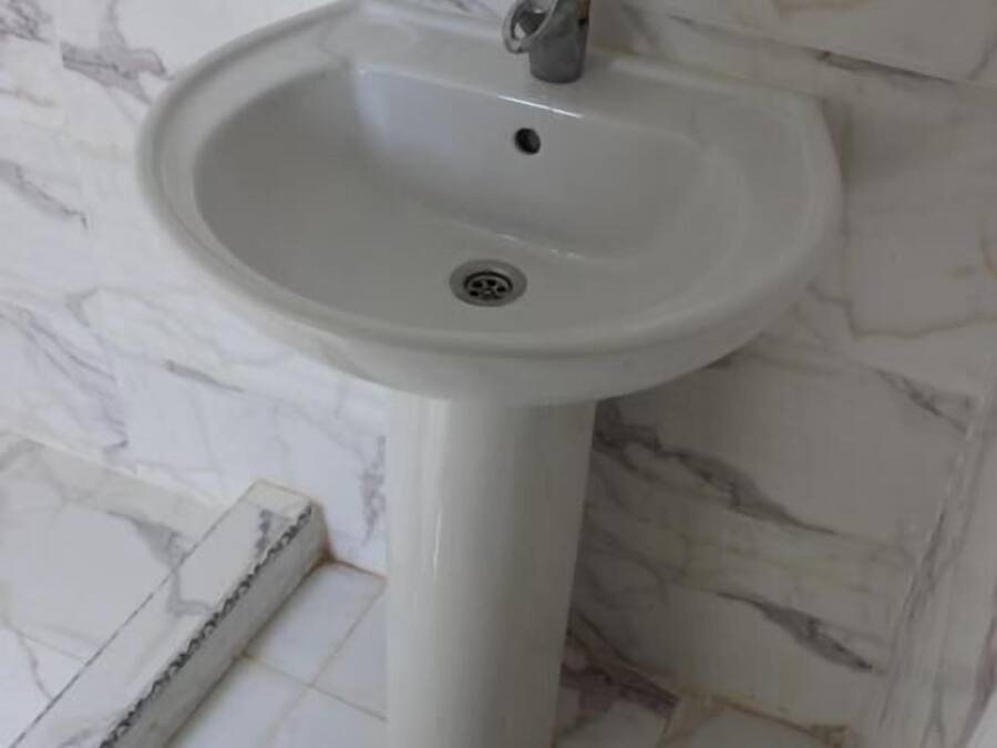 A bathroom with a white pedestal sink, marble walls, and white tiled flooring.