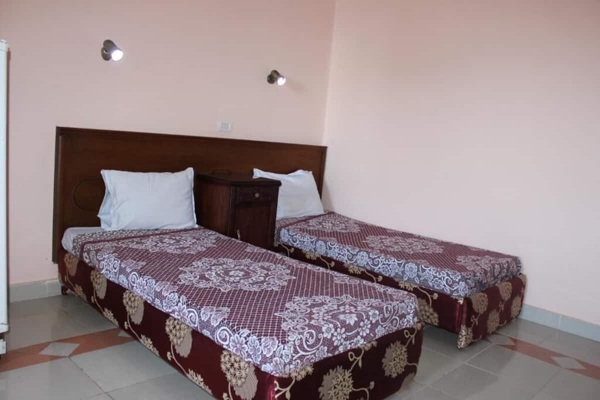 Ein Zimmer mit zwei Einzelbetten, jeweils mit einem gemusterten Überwurf und weißen Kissen. Zwei Wandleuchten und ein kleiner Nachttisch mit Lampe sind vorhanden.