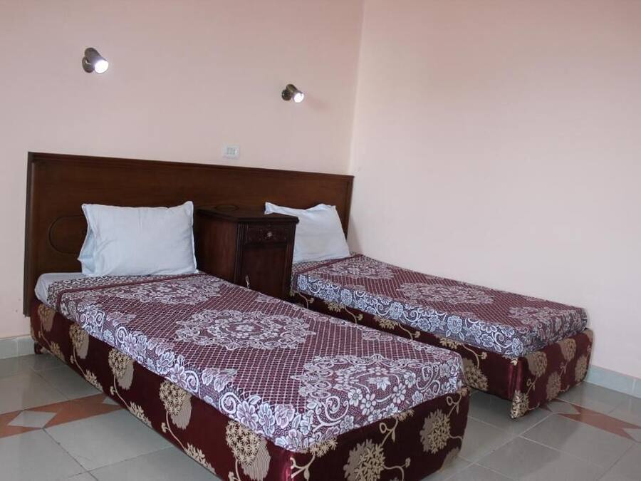 A room with two single beds, each with a patterned bedspread and white pillows. There are two wall-mounted lights and a small bedside table with a lamp.