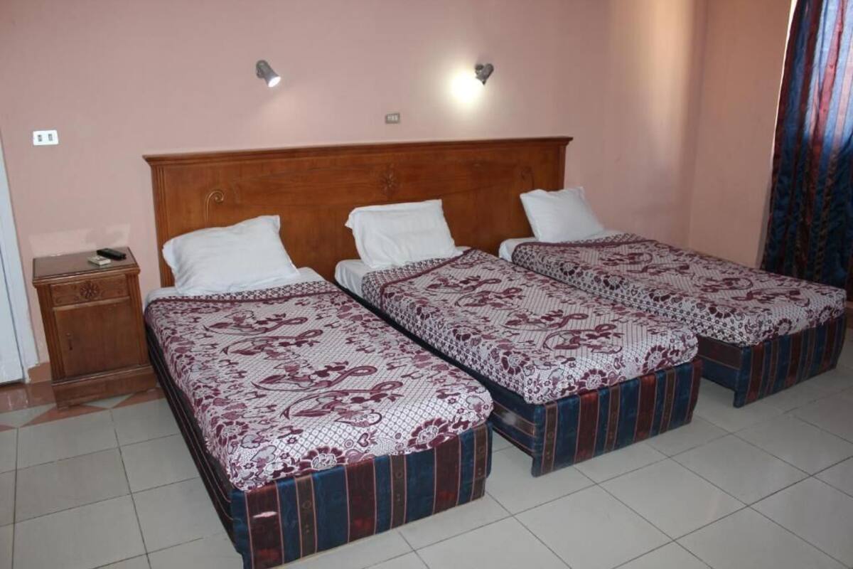 Ein Hotelzimmer mit drei Einzelbetten, jeweils mit einem gemusterten Überwurf und einem gestreiften Fußteil. Ein Holznachttisch mit Lampe und Fernbedienung steht daneben. Die Wände sind rosa und es gibt ein Fenster mit Vorhängen.