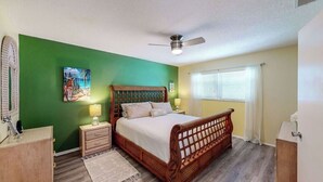 Une chambre avec un lit en bois, des murs verts, un ventilateur de plafond, une fenêtre avec des stores et une table de chevet avec une lampe.
