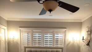 Ein Schlafzimmer mit einem gebuschten Kopfteil, einem Bett mit einer gesteppten Bettdecke, einem Nachttisch, einer Stehlampe und einem Deckenventilator.