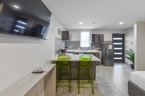 A modern kitchen with a flat-screen TV, two bar stools, a refrigerator, and a window.