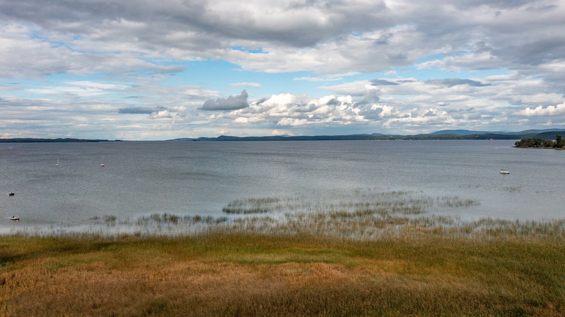 Vista amplia de un lago con barcos, orillas cubiertas de hierba y colinas lejanas bajo un cielo nublado.