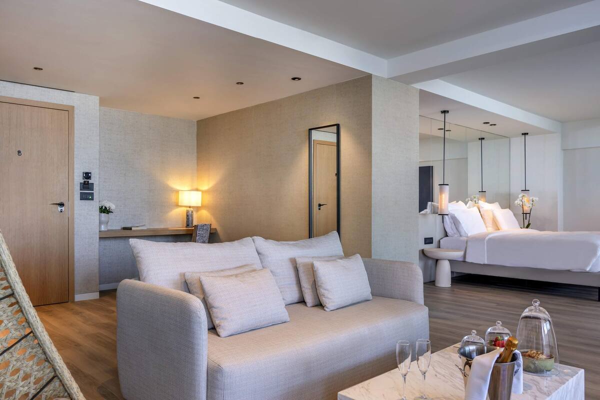 Ein modernes Hotelzimmer mit einer Couch, einem Bett, einem kleinen Tisch mit Gläsern und einer Ablage sowie einem Spiegel.