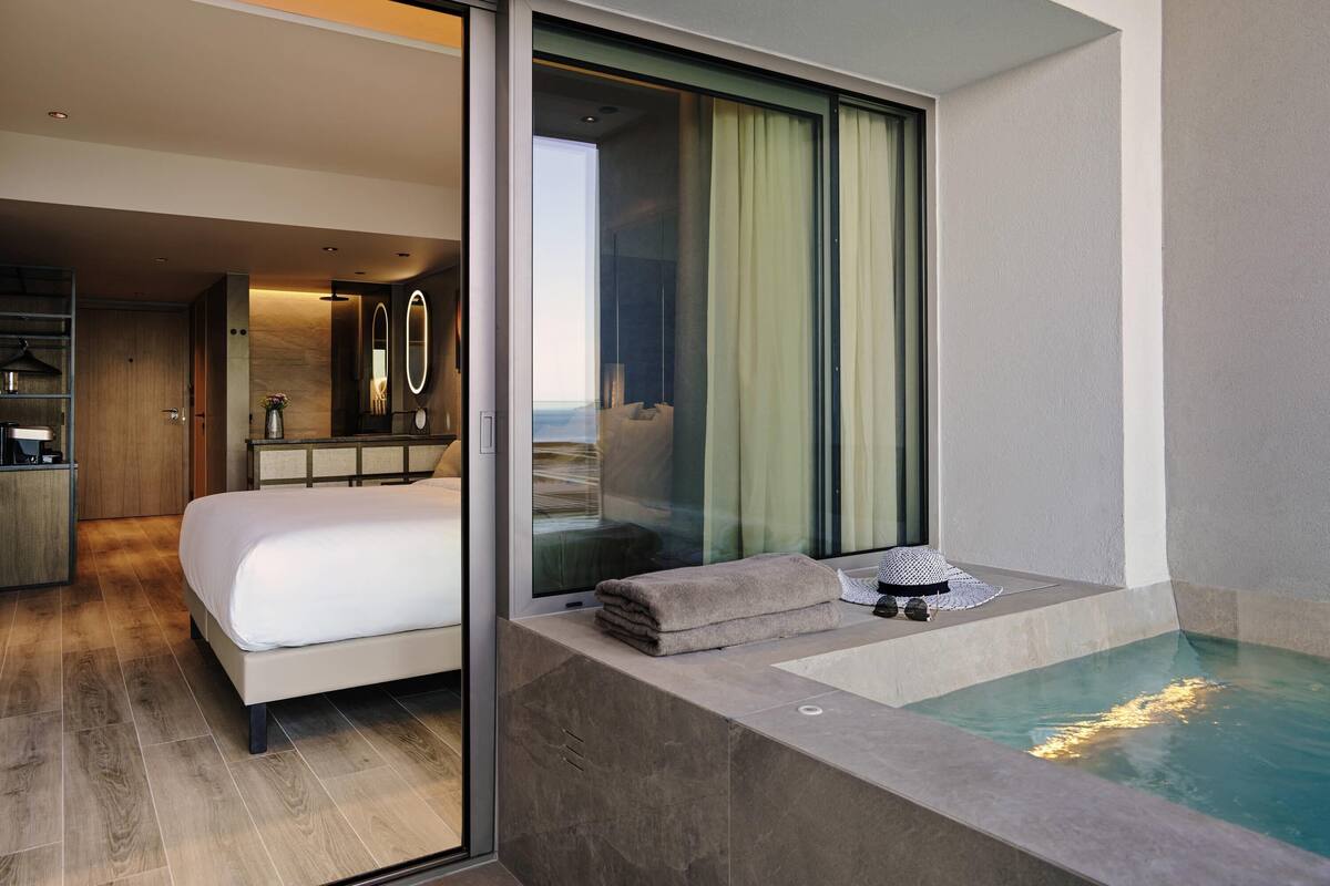 Ein modernes Hotelzimmer mit einem großen Bett, einer Glastür, die auf einen Balkon mit Aussicht führt, einer kleinen Sitzecke mit Whirlpool und einem Badezimmer mit Spiegel und Waschtisch.