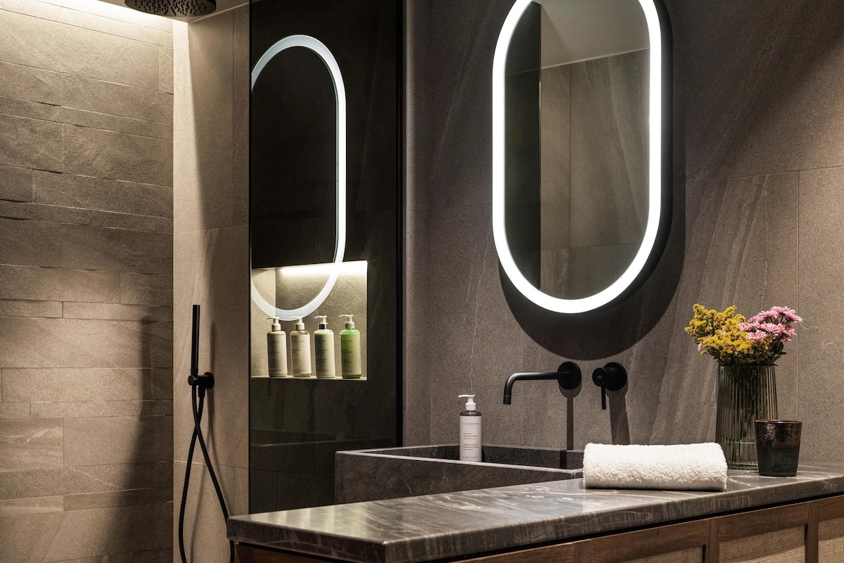 A modern bathroom with a large mirror, a showerhead, a sink, and toiletries.