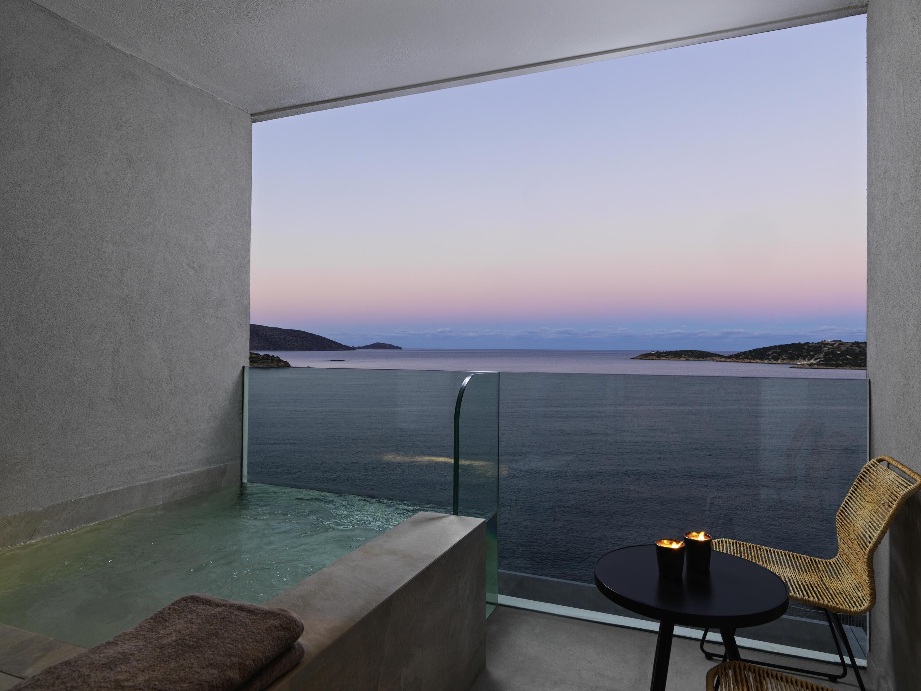 A balcony with a glass barrier, a hot tub, a wicker chair, and a table with a candle.