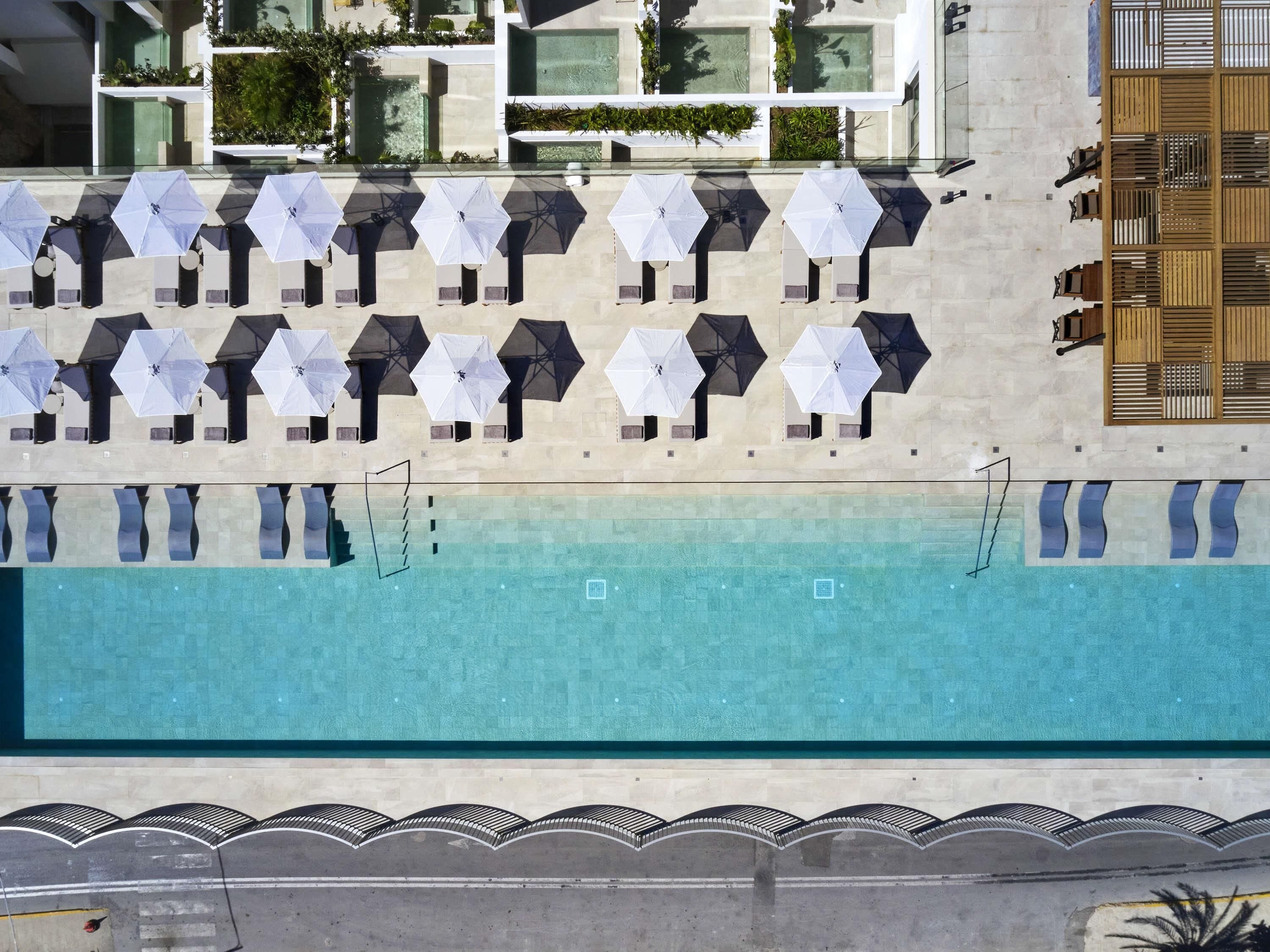 Una piscina en la azotea con múltiples sombrillas y sillones reclinables, rodeada por un edificio moderno con terrazas y vegetación.