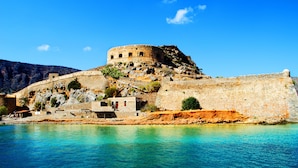 A historic stone fortress with a round tower, situated on a rocky hillside overlooking a clear blue sea.