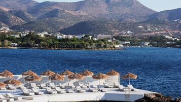 A coastal resort with a pool area featuring sun loungers and thatched umbrellas, overlooking a calm sea and a mountainous backdrop.