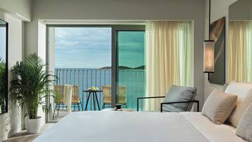 A modern hotel room with a large bed, a balcony with a view of the sea, and a dining area with a table and chairs.