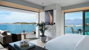 A modern hotel room with a large bed, a seating area with a table, a potted plant, and a view of the sea and mountains.