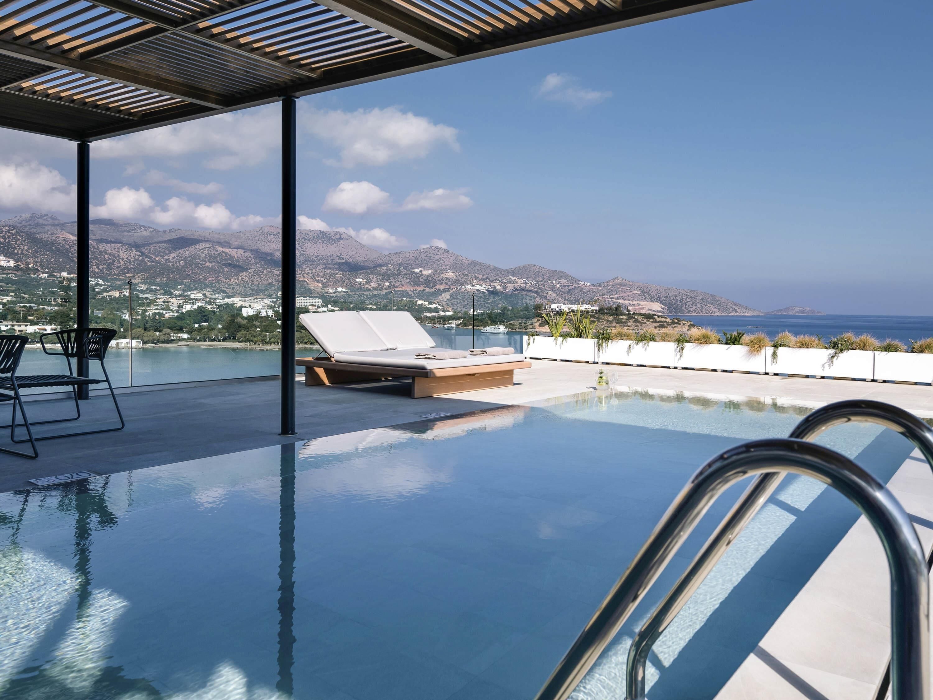 Una piscina infinita con una silla de descanso y una mesa, cielo despejado y un paisaje montañoso al fondo.