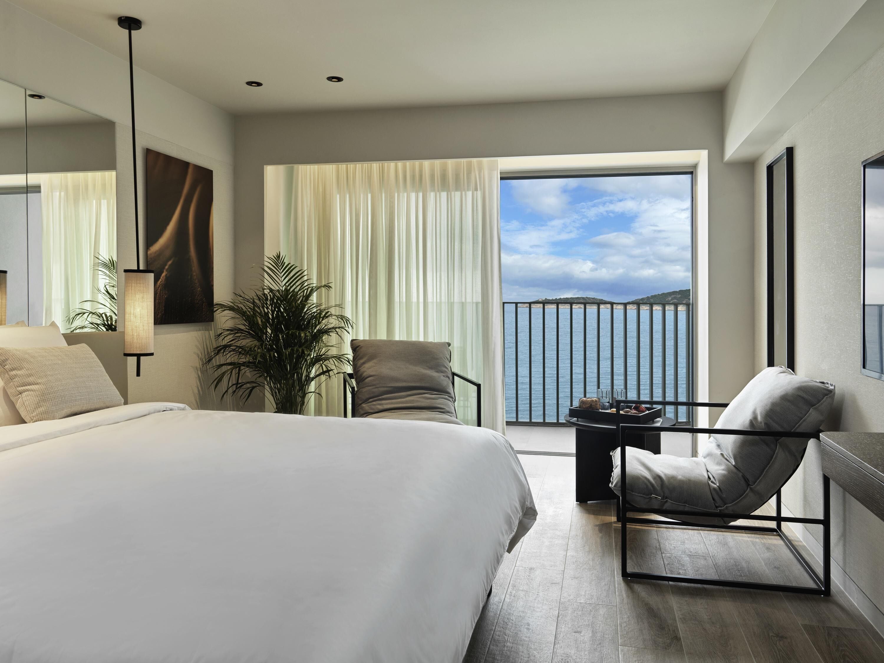 A modern hotel room with a large bed, a seating area with a view of the sea, and a balcony with a railing.