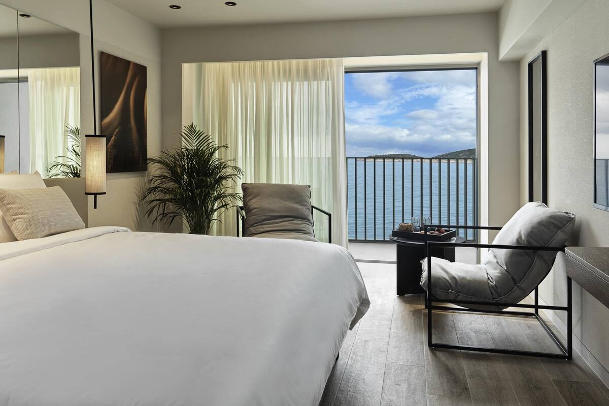 Ein modernes Hotelzimmer mit einem großen Bett, einer Sitzecke mit Meerblick und einem Balkon mit Geländer.