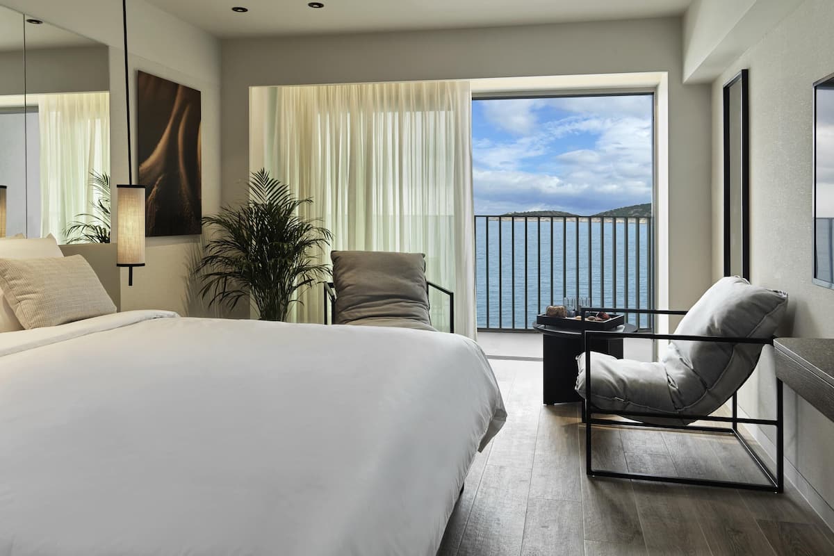 A modern hotel room with a large bed, a seating area with a view of the sea, and a balcony with a railing.
