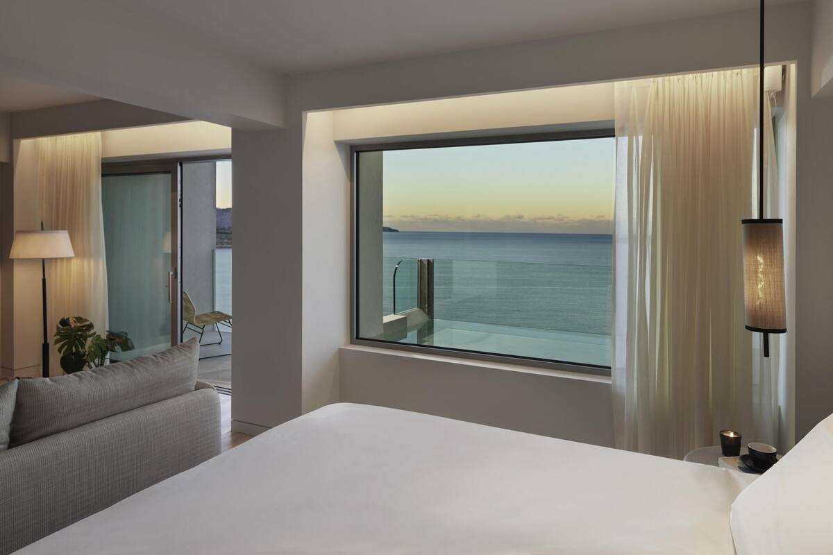 Ein Hotelzimmer mit großem Fenster und Meerblick, einem Bett, einem Sessel, einer Lampe und einem kleinen Tisch mit einer Tasse.