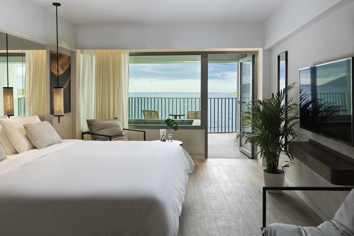 Ein geräumiges Schlafzimmer mit einem großen Bett, einem Balkon mit Meerblick und einem an der Wand befestigten Flachbildfernseher.