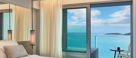 A modern hotel room with a large bed, a balcony with a view of the sea, and a small table with chairs.