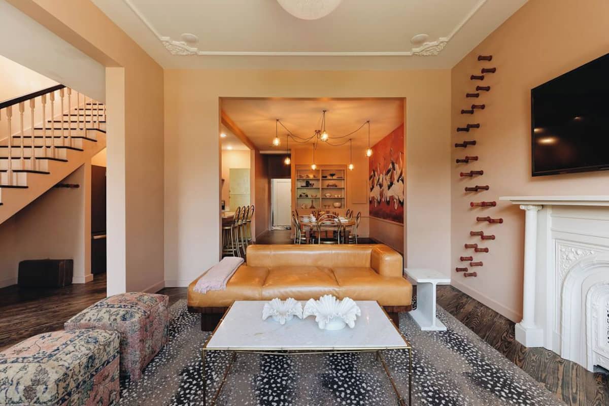 A spacious living room with a leather sofa, a coffee table, and a fireplace. A staircase is visible on the left, and a dining area is in the background.